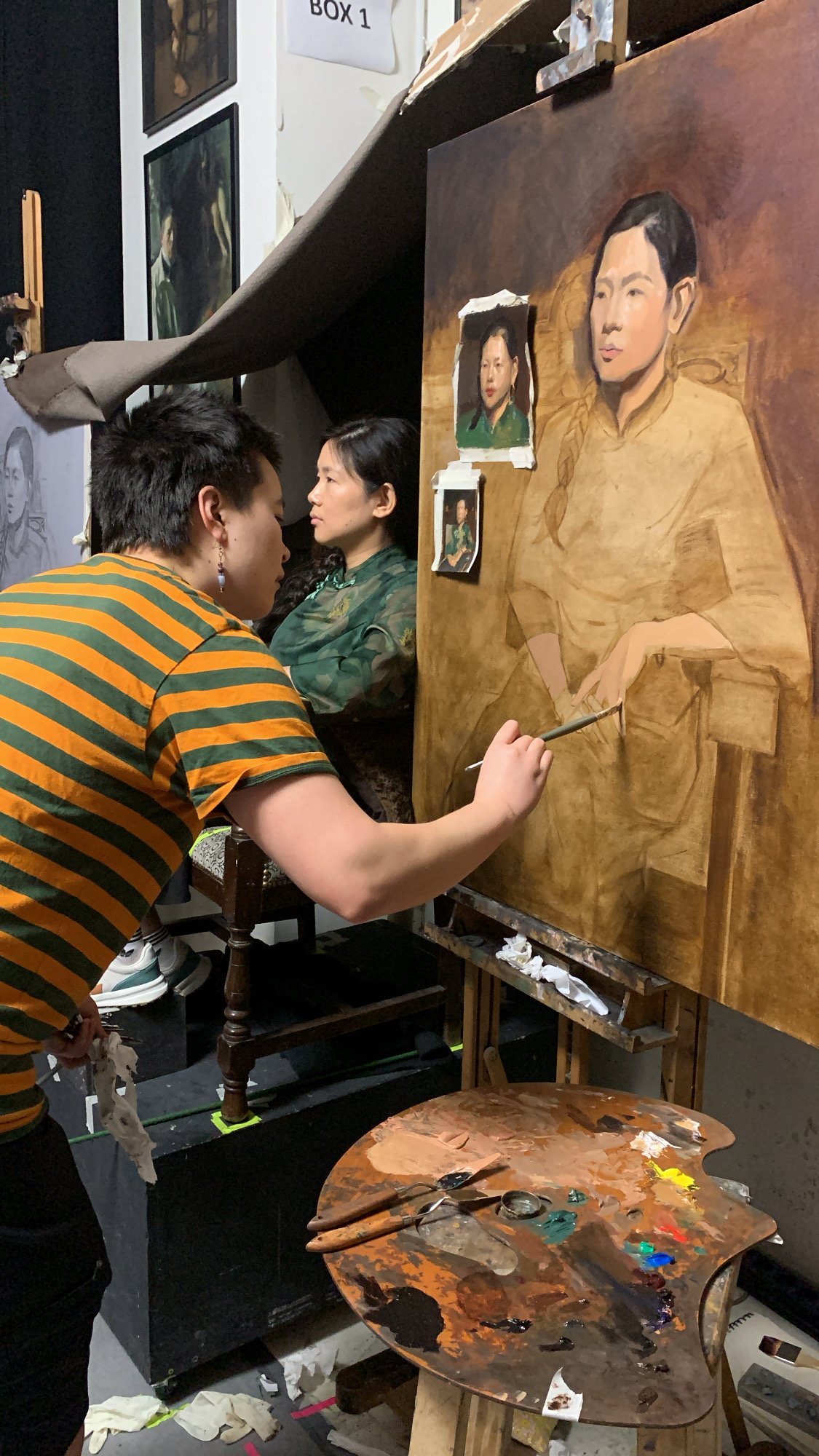 T. painting the large portrait canvas, the warm initial layer in progress with Jie seated in the background